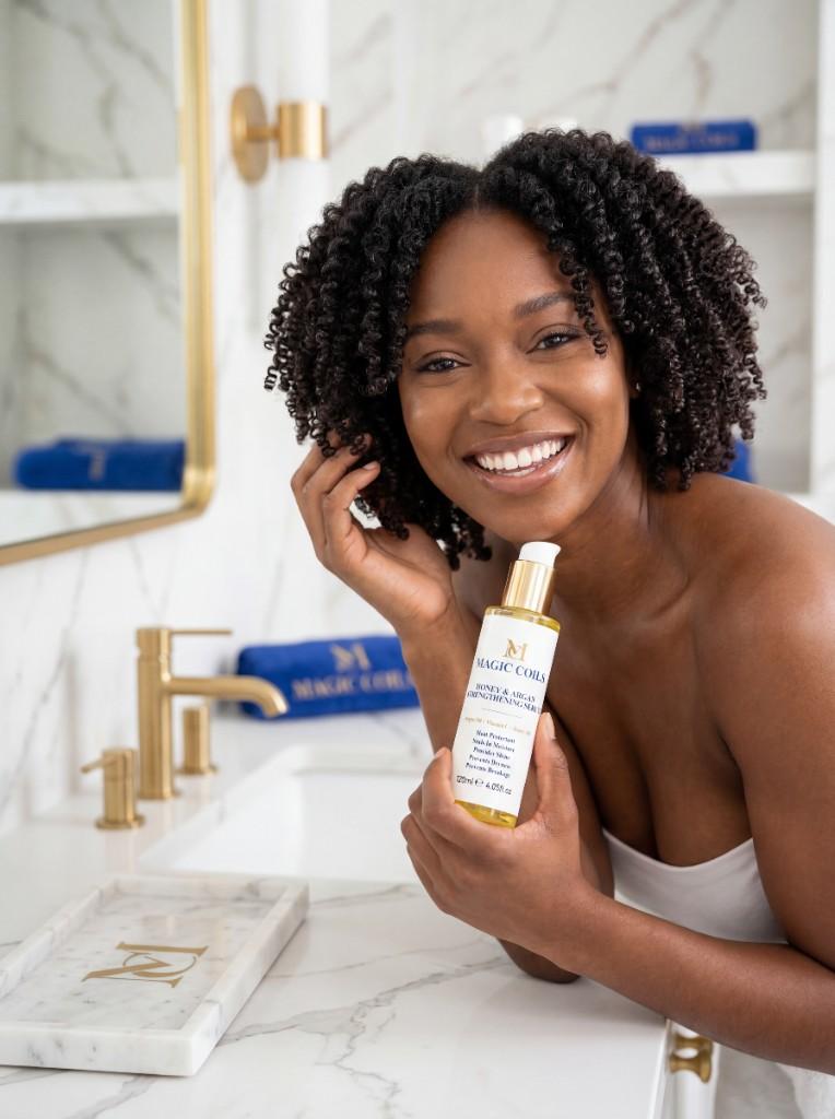 Smiling Black woman with natural hair holding Magic Coils Strengthening Serum
